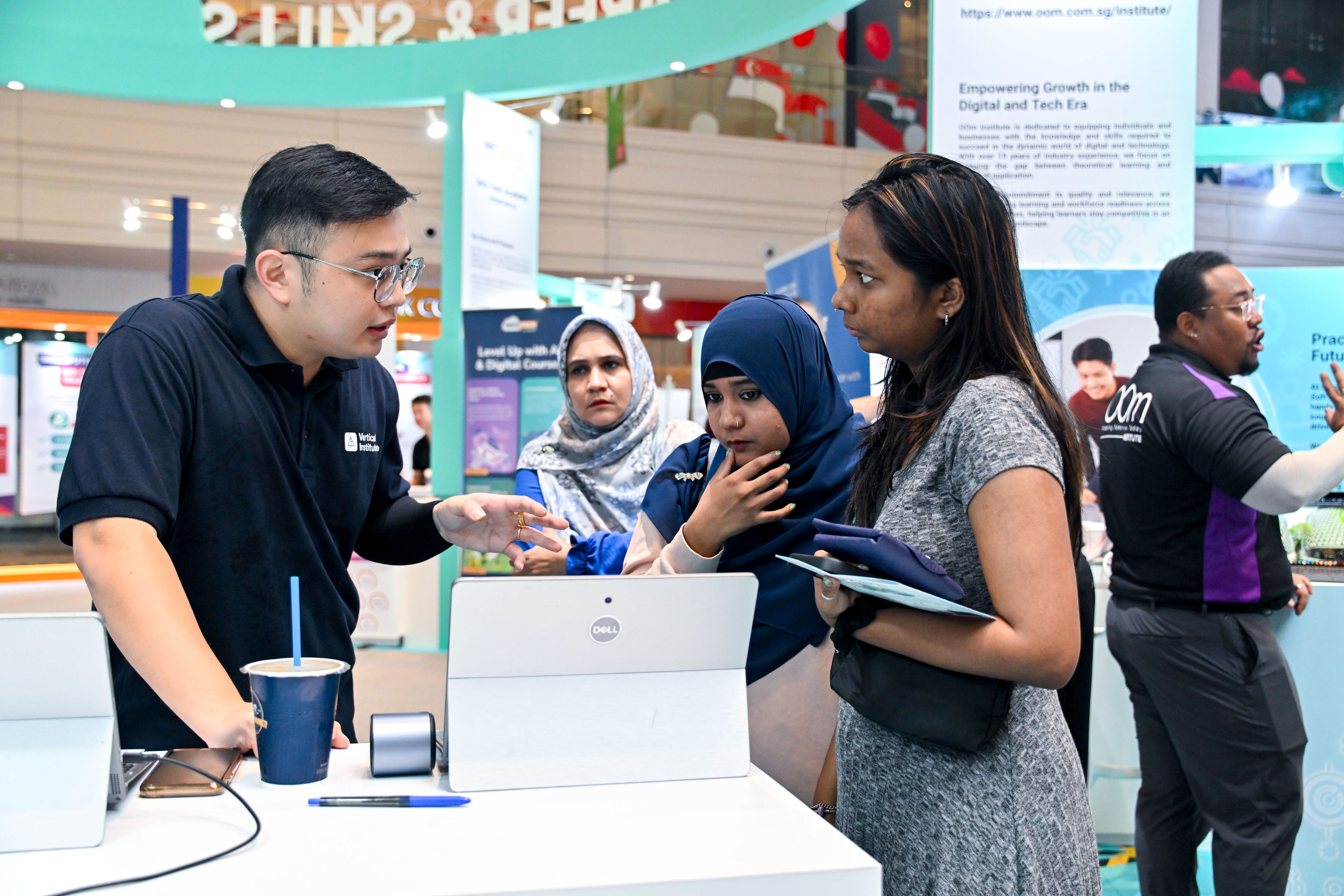 Three women speaking with a representative from the Verticle Institute at an exhibition booth about training opportunities